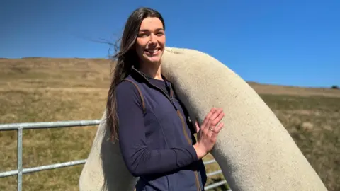 BBC A young woman with long brown hair. She has a wool log over one shoulder and is standing in front of grassland. The sky behind her is bright blue.
