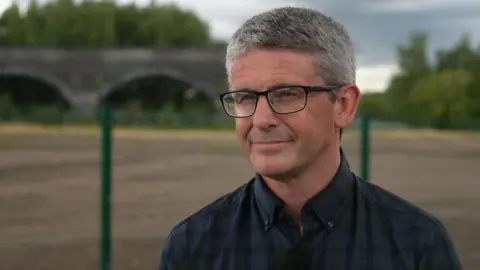 Gavin White has short hair and is wearing a dark blue shirt and dark framed glasses. He is stood in front of a green metal fence with open area of brownfield and a railway was bridge in the background.