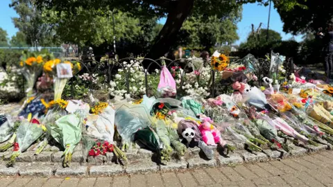 Essex Police Many flowers and soft toys have been left in a circle in the park where Leonna died. Trees are in the distance. The sun shines on the toys and floral bouquets.