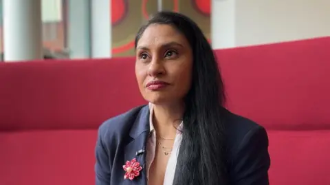 A woman with long dark hair sitting on a red sofa. She is wearing a white shirt, navy blazer, and as a red button brooch.