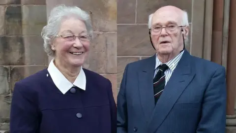 A older man and woman look into the camera. The woman is wearing a dark purple coat with a white collar and is smiling. The man is wearing a navy suit, striped shirt and striped tie. They are both wearing glasses.
