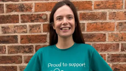 A woman with long brown hair smiles while standing in front of a brick wall. She is wearing a green t-shirt which says "proud to support" on it.