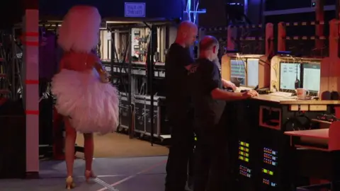 Shaun Whitmore/BBC The back of a performer dressed in a white feather head piece carrying feather fans under her arm. Two men dressed in black are looking away from the camera who are working backstage with computer monitors in front of them on a desk.