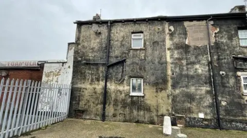 LDRS The side of an old looking house. Plaster is peeling from the walls and it has what appears to be dark mould, or just lots of dirt, all over. There are two small windows and a metal grate fence is to the left of it. 