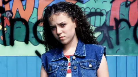 Dani Harmer as Tracy Beaker in the BBC adaptation of the books. She is wearing a stripy red and white t-shirt and a denim waistcoat.