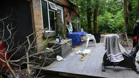 Devon and Cornwall Police Side view of a wooden cabin with white UPVC windows and door and wood decking in front. The deckin is clutter with an exercise bike which has a towel hung on it, a broom, chairs, a ghille suit hanging from the cabin, a paint can and other items. Green forest can be seen in the background.