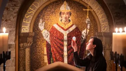 A view across a stone tomb, lined by large candles, towards a mural of St William. He is depicted wearing a red robe and holding a gold cross. A woman stands beside the tomb and holds up an artefact. 
