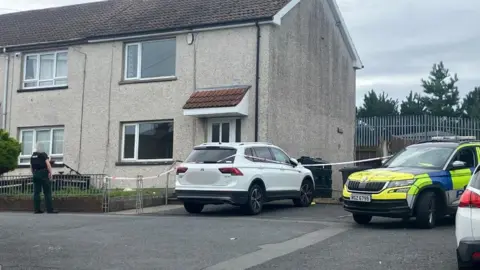 A police car sits outside a cream coloured house. There is red and white police tape cordoning off the scene. 