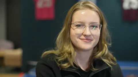 Jamie Niblock/BBC Bethan Manning sitting in an office. She has light brown shoulder-length wavy hair and is wearing clear framed plastic glasses and a black zipped top. 