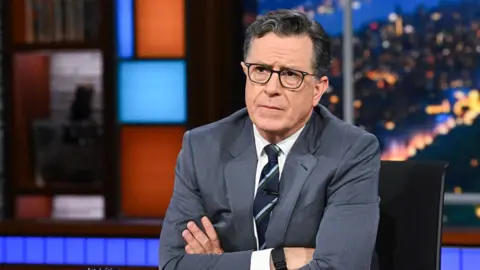Stephen Colbert sitting at a desk with his arms crossed frowning. 
