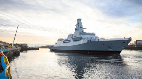 PA Media A large grey warship arriving at a shipyard on a river