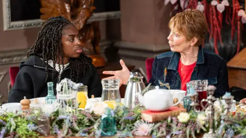 Cat Burns and Celie Imrie sitting next to each other and talking at the breakfast table in The Traitors