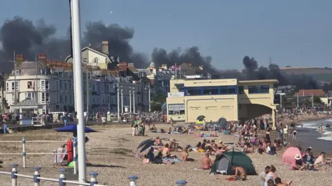 A view of Weymouth beach with crowds of people enjoying the sunshine, with thick black smoke billowing in the background