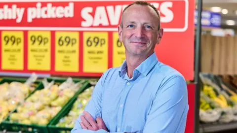 Giles Hurley, Aldi UK chief executive, poses with his arms folded in front of the fruit and veg aisle in an Aldi supermarket