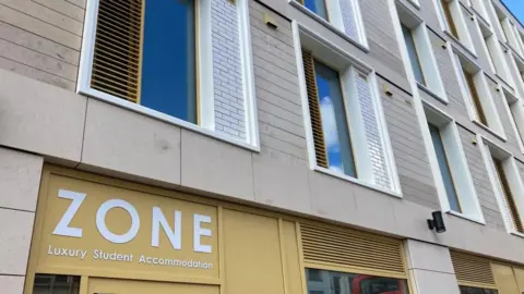 Close-up view of the exterior of a block of flats with several storeys. The building is made up of grey bricks with narrow windows, which have white frames and wooden louvres.  The ground floor has some yellow facing, on which is written "Zone Luxury Student Accommodation".