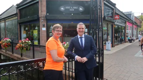 SOSE Two people, a woman in an orange top, and a man in a blue suit, lean against railings near a high street