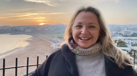 Katya Maddison wearing a grey jumper and patterned scarf with a beach in the background. She has long blonde hair and is smiling directly at the camera.