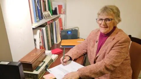 Plymouth City Council Linda Gilroy sits at a desk with a pencil in her hand. She is wearing a pink blazer and looks at the camera.