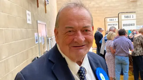 Conservative Councillor Adrian Hardman is smiling at the camera, wearing a blue suit and a blue Conservative rosette.