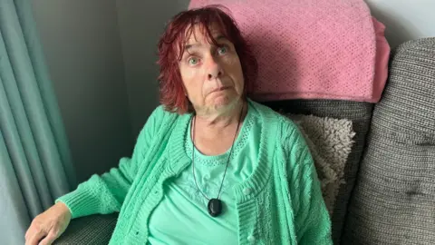 Woman in green top and cardigan with short red hair sat down on sofa 