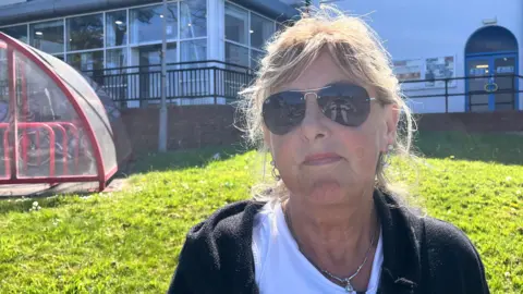 A woman with blond hair and dark sunglasses stands in front of a leisure centre building. She is wearing a black cardigan, white T-shirt and silver necklace. Behind her is a green lawn and red-framed glass bike shelter.