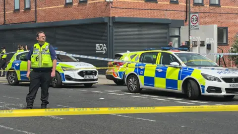 Police cordon at the scene. Three police cars are parked behind yellow tape while an officer stands in the road