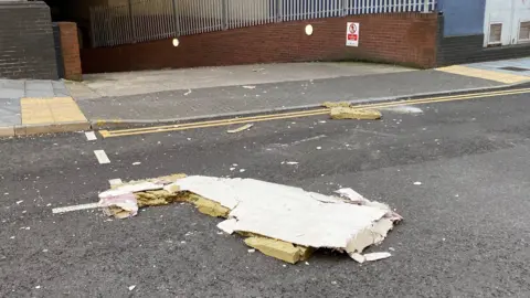 Bits of drywall are scattered around on a road with an entrance to a car park seen in the background.