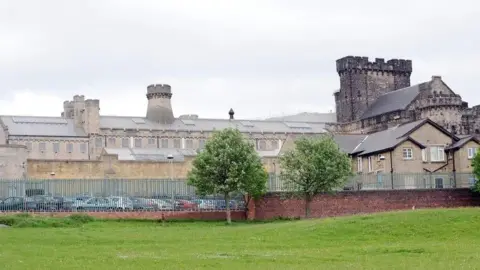 A view of HMP Leeds from outside its boundary wall and fence. The prison estate is a mixture of modern light brown buildings and the remains of what appears to be a castle.