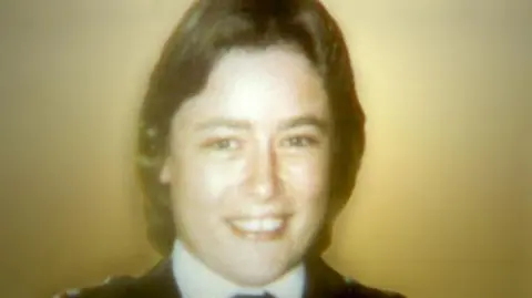 Official police photo of Yvonne Fletcher wearing a police uniform in front of a yellowish wall. She has short hair and is smiling at the camera. The picture was taken in 1984 and is fairly low-resolution.