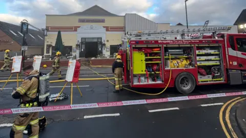 Four firefighters are on the left and in the middle of the photo. A fire engine is to the right and a 'fire do not cross' cordon is in the foreground. A mainly yellow building is in the background.