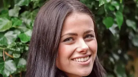 India Chipchase is smiling and looking at the camera. She has long dark hair and there is a green bush behind her. 