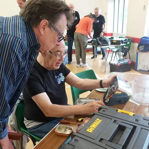 Lichfield Repair and Share Cafe A man sits next to some tool boxes on a table, while another man leans over and looks at what he is doing.