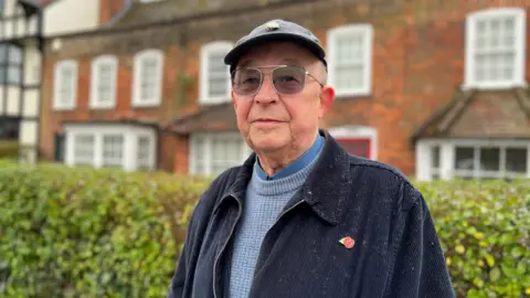 A man wearing a blue corduroy jacket and a cap, looking straight at the camera. There's a hedge and a brick house behind him.