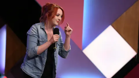 A woman standing on the left with red hair tied up in a ponytail wearing a blue denim shirt, who is holding a black microphone and smiling