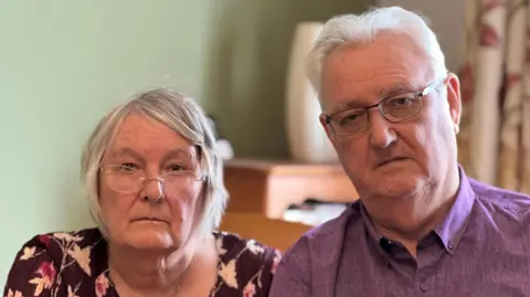 Katie and Peter McCombe are pictured looking disappointed and annoyed in the dining room of their home. They both have grey hair and are wearing glasses. The walls behind them are green and they have flowered curtains to their left. There is cabinet blurred in the background with a vase on top. Mr McCombe is wearing a mauve shirt. His wife is wearing a black and floral top.