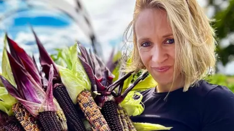 Lucy Hutchings smiles at the camera. She holds a number of colourful corn cobs. She has blonde hair and a navy t-shirt.