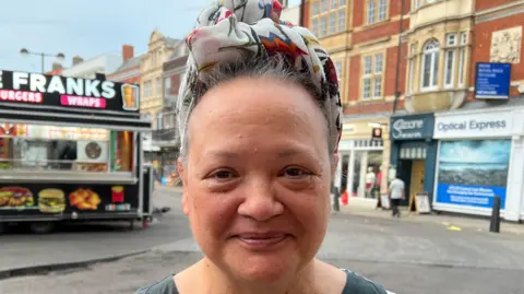 Mousumi Bakshi/BBC A woman - Shareena Howley - looking straight at the camera with a neutral expression. She is wearing a black sleeveless T-shirt and is standing in front of a row of shops and a takeaway food outlet.