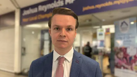 The MP for Maidenhead Joshua Reynolds stands outside the front of his office. He is wearing a blue suit with a pink tie and he has a serious look on his face. 