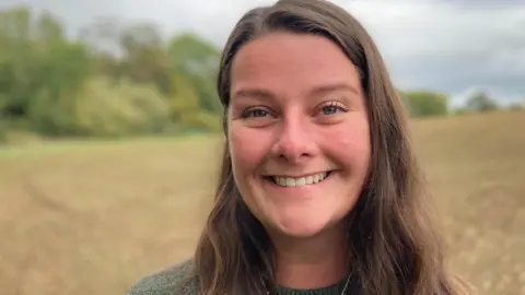 A woman wearing a green jumper and silver necklace, standing in a field, with brown hair and smiling