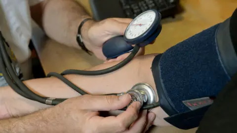 A doctor is using a stethoscope and blood pressure monitor on the arm of a patient. The image is a close-up of the patient's forearm and the doctor's hands.