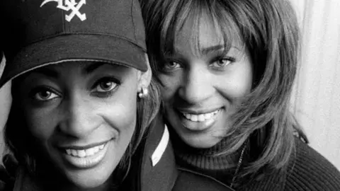 A close cropped black and white image of two women's faces. They are both looking at the camera and smiling, the woman on the left is wearing a baseball cap, the woman on the right has should length hair.