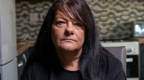 Yeti Television/BBC Cymru Wales Bethan Callaghan's mother, Gaynor. She has black long hair and a side fringe. She has brown eyes and stares blankly into the camera. She sits at the forefront of the photo and a blurred kitchen with black and white tiles can be seen in the background. 