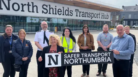 A group of nine people standing in front of a large glass-fronted building which has a large sign reading 'North Shields Transport Hub' on the side. The group is made up of six women and three men. Susan Dungworth is standing in the middle of the photo and has light red, curly hair. She is holding one side of a black and white banner which reads 'Safer Transport Northumbria'. She is wearing a black jacket and red floral dress. A bald man is holding the other side of the banner and is wearing a blue shirt and black trousers.