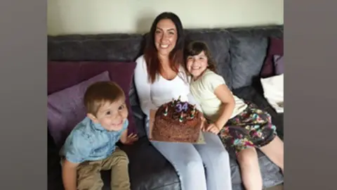 Jade Horton A boy in a blue shirt sits on a sofa next to a woman in a white top and a girl in a yellow top, both holding a birthday cake.