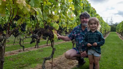 David Llewellyn David Llewellyn who has salt and pepper hair and a beard with his son who has longish blond hair beside grape vines
