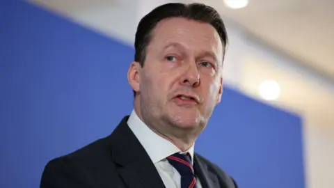 PA Media Russell Findlay, who has dark hair swept back, speaks in front of a blue background. He is wearing a business suit 