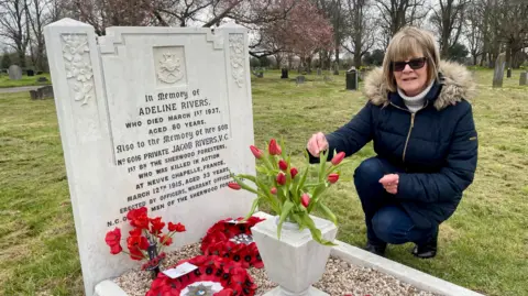 BBC A white headstone, grave and outdoor vase in a graveyard. A women crouches beside the grave putting red tulips into the vase. She has short fair hair, sunglasses, a navy coat, dark blue jeans and black boots. There are two poppy wreaths and a bunch of poppies laid on the gravel of the grave. There are trees and other darker graves in the background. The headstone is engraved with flowers, a cross and words. Those words read. "In memory of Adeline Rivers who died March 1st 1937 aged 80 years. Also to the memory of her son, No. 6016 Private Jacob Rivers, V.C. 1st Bt The Sherwood Foresters, who was killed in action at Neuve Chapelle, France, March 12th 1915, aged 33 years. Erected by officers, warrant officers, N.C.O. and men of the Sherwood Foresters." 