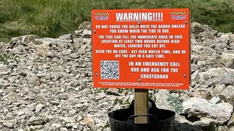 Bay Search and Rescue Team A red warning sign telling people to call 999 in an emergency and to scan QR codes to find out tide times..