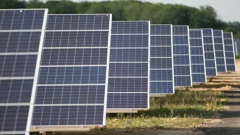 An array of solar panels. There are nine in the picture, with some grass on the ground and trees in the background.