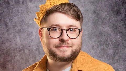 Dash Silva A man with glasses and neatly styled beard wears a yellow corduroy jacket and a knitted crown in a matching colour. He's in front of a marble-effect studio background in this headshot-style photo 